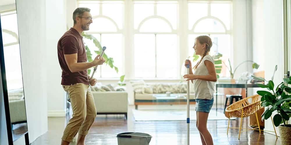 A father and daughter share a laugh while mopping the wooden floor in a spacious living room.